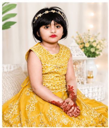 Young girl wearing a yellow dress with floral embroidery and a headband, sitting indoors.